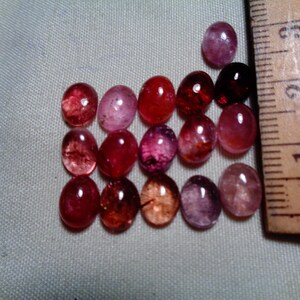 Puede incluir: Una colección de gemas ovaladas en tonos rojos, rosas y morados. Una regla de madera con marcas de centímetros se coloca junto a las gemas. Las gemas varían en tamaño, la más grande mide aproximadamente 2,5 cm.