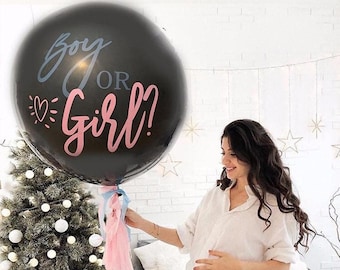May include: A large black balloon with the text "Boy or Girl?" in blue and pink script. The balloon is decorated with pink and blue tassels. The balloon is held by a person in a white shirt.