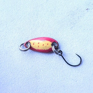 May include: A fishing lure with a red and yellow painted oval body, two silver rings, and a black hook. The lure is designed to attract fish.