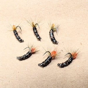 May include: Six handmade fishing flies. Each fly features a dark body with a silver bead, a curved hook, and a tuft of orange and tan fibers. The flies are arranged on a light brown surface.