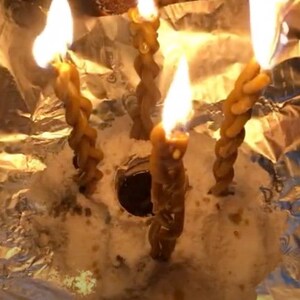 May include: Four brown candles are burning in a silver foil container. The candles are twisted and have a rough texture. The flames are bright and orange.