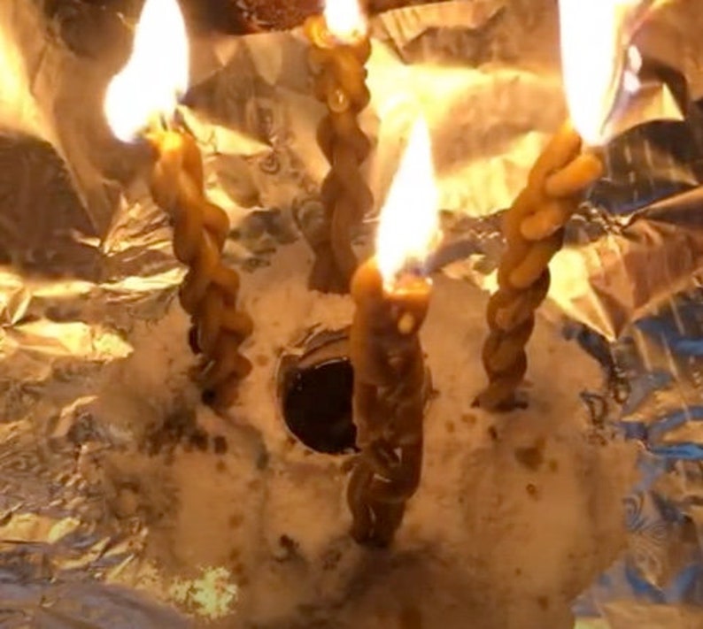 May include: Four brown candles are burning in a silver foil container. The candles are twisted and have a rough texture. The flames are bright and orange.