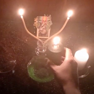May include: A candle holder with three lit candles, two with long, thin, wooden wicks and one with a shorter, white wick. The candles are arranged in a triangular shape. The candle holder is made of clear glass and has a decorative design. There is a small, green, circular object in the foreground.