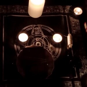 May include: A black cloth with a white pentagram design and three lit candles. The candles are in glass holders and are placed on the cloth.