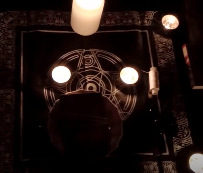 May include: A black cloth with a white pentagram design and three lit candles. The candles are in glass holders and are placed on the cloth.