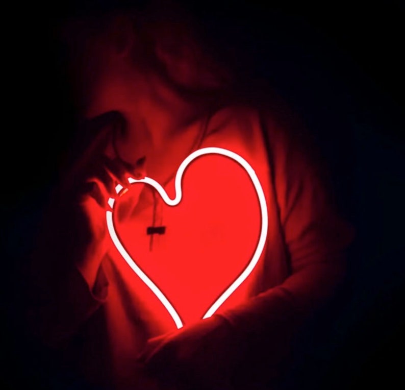 May include: A person holding a neon red heart-shaped sign in front of their chest. The sign is glowing brightly against a dark background.