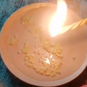 May include: A close-up of a lit candle with a yellow wick. The candle is in a clear glass container and the wax is melted and has a light brown color.