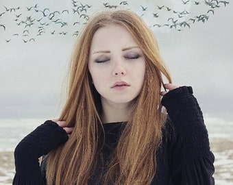 Op de afbeelding: Een jonge vrouw met lang, steil, roodbruin haar draagt een zwart shirt met lange mouwen. Ze heeft haar ogen gesloten en kijkt naar beneden. De achtergrond is een wazig beeld van een besneeuwd landschap met een zwerm vogels die erboven vliegen.