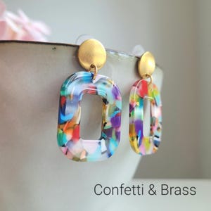 K&ouml;nnte beinhalten: Ein Paar farbenfrohe, offene rechteckige Ohrringe mit einem Konfetti-&auml;hnlichen Design. Die Ohrringe haben einen goldfarbenen Stecker und einen Messingfarbenen Akzent. Der Text "Confetti & Brass" befindet sich am unteren Bildrand.