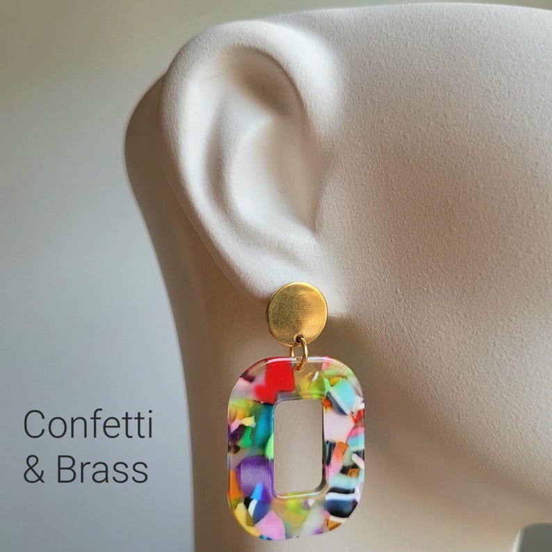 K&ouml;nnte beinhalten: Ein Paar bunte Konfetti-Ohrringe mit einem Messing-Stecker und einem rechteckigen Design mit offenem Zentrum. Die Ohrringe weisen eine lebendige Mischung aus roten, blauen, gr&uuml;nen und violetten Farben auf. Der Text "Confetti & Brass" ist sichtbar.