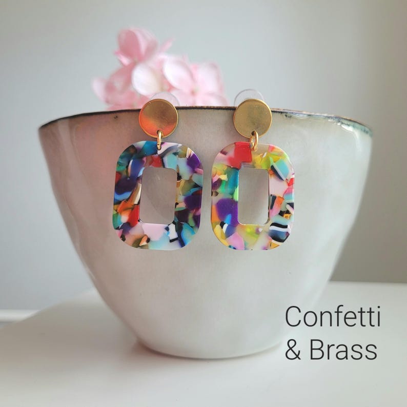 K&ouml;nnte beinhalten: Rechteckige Ohrringe mit Konfetti-Design und Messingdetails. Die Ohrringe haben ein farbenfrohes Muster und eine offene Mitte. Der Text "Confetti & Brass" ist im Bild zu sehen. Sie werden vor einer wei&szlig;en Schale mit einer rosa Blume pr&auml;sentiert.