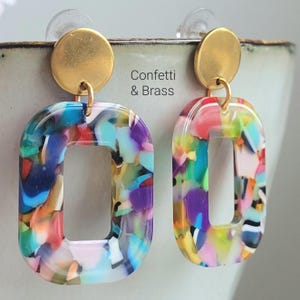 K&ouml;nnte beinhalten: Ein Paar bunte, rechteckige Ohrringe mit einem Konfetti-&auml;hnlichen Design. Die Ohrringe haben einen goldfarbenen kreisf&ouml;rmigen Stecker und einen passenden goldfarbenen Verbindungsring. Der Text "Confetti & Brass" ist sichtbar.