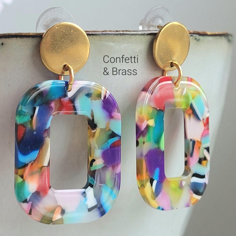 K&ouml;nnte beinhalten: Ein Paar bunte, rechteckige Ohrringe mit einem Konfetti-&auml;hnlichen Design. Die Ohrringe haben einen goldfarbenen kreisf&ouml;rmigen Stecker und einen passenden goldfarbenen Verbindungsring. Der Text "Confetti & Brass" ist sichtbar.
