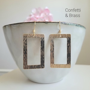 Könnte beinhalten: Ein Paar goldfarbene, rechteckige Ohrringe mit strukturierter Oberfläche. Die Ohrringe hängen vor einer weißen Schale, im Hintergrund eine rosa Blume. Der Text "Confetti & Brass" ist sichtbar.