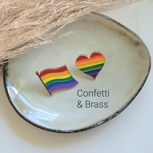 Könnte beinhalten: Zwei Emaille-Pins auf einem weißen Keramikteller. Ein Pin ist eine Regenbogenflagge und der andere ein Regenbogenherz. Der Text "Confetti & Brass" ist unter den Pins gedruckt.
