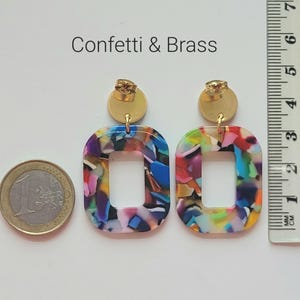K&ouml;nnte beinhalten: Bunte, rechteckige Ohrringe mit einem Konfetti-Muster und goldfarbenen Pfosten. Die Ohrringe haben eine hohle Mitte und werden mit einem Lineal mit Zentimetermessungen gezeigt. Der Text "Confetti & Brass" befindet sich &uuml;ber den Ohrringen.