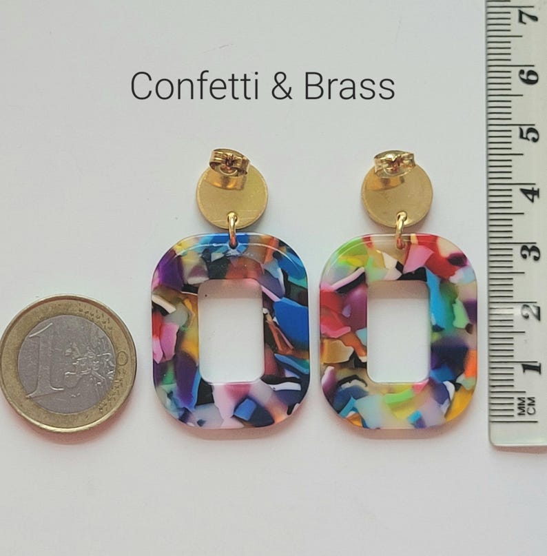 K&ouml;nnte beinhalten: Bunte, rechteckige Ohrringe mit einem Konfetti-Muster und goldfarbenen Pfosten. Die Ohrringe haben eine hohle Mitte und werden mit einem Lineal mit Zentimetermessungen gezeigt. Der Text "Confetti & Brass" befindet sich &uuml;ber den Ohrringen.