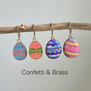 Könnte beinhalten: Vier bunte Ostereier-Ohrringe hängen an einem Ast. Jeder Ohrring hat ein einzigartiges Design mit blauen, rosa, grünen, lila und gelben Farbtönen. Die Ohrringe haben goldfarbene Haken. Der Text "Confetti & Brass" steht darunter.