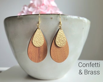 Puede incluir: Un par de pendientes en forma de lágrima con una parte superior de metal martillado dorado y una parte inferior de madera marrón. Los pendientes cuelgan de un cuenco blanco. El texto "Confetti & Brass" es visible debajo de los pendientes.