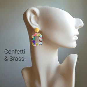 K&ouml;nnte beinhalten: Confetti & Brass Ohrringe. Diese Ohrringe haben eine rechteckige Form mit einem bunten Konfetti-Design. Sie sind an einem kleinen, runden, goldfarbenen Stecker befestigt. Der Text "Confetti & Brass" ist sichtbar.