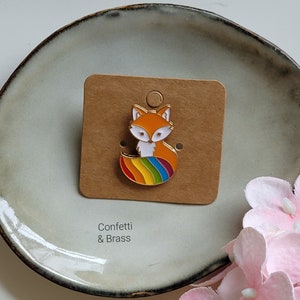 Könnte beinhalten: Eine Regenbogen-Fuchs-Emaille-Anstecknadel auf einer braunen Karte mit dem Text "Confetti & Brass".