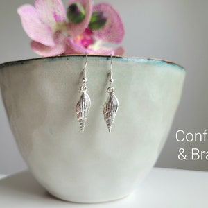 Könnte beinhalten: Silberne Muschel-Ohrringe mit einer strukturierten Oberfläche. Die Ohrringe hängen an einer weißen Schale mit einer rosa Orchidee im Hintergrund. Der Text "Confetti & Brass" ist in der unteren rechten Ecke des Bildes sichtbar.