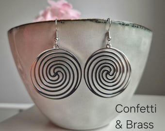 Puede incluir: Un par de pendientes en espiral plateados colgando frente a un cuenco de color claro con una flor rosa. Los pendientes tienen un diseño circular con espirales concéntricas. El texto de la imagen dice "Confetti & Brass."