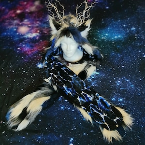 Puede incluir: Una bufanda con capucha de piel sintética blanca y azul con una bufanda estampada en negro y azul unida. La capucha tiene un diseño de piel sintética blanca y azul con un par de orejas blancas y un diseño blanco en forma de rama en la parte superior. La bufanda está fotografiada sobre un fondo azul oscuro con estrellas blancas y rosas.