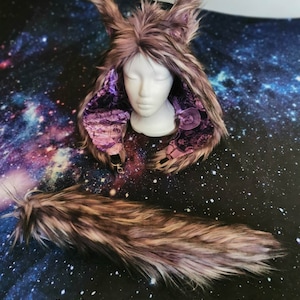 Puede incluir: Un disfraz de capucha de color gris y morado con una cola a juego. La capucha tiene orejas grandes y peludas y un forro morado. La cola es larga y esponjosa.