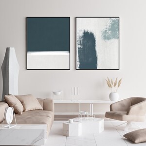 May include: Two abstract paintings in black frames, one with a teal blue and white horizontal stripe design, the other with a teal blue brushstroke on a white background. The paintings are hanging on a white wall above a beige sofa and a white coffee table.