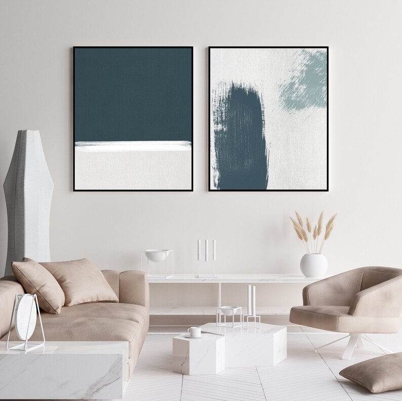 May include: Two abstract paintings in black frames, one with a teal blue and white horizontal stripe design, the other with a teal blue brushstroke on a white background. The paintings are hanging on a white wall above a beige sofa and a white coffee table.