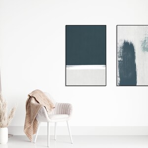 May include: Two framed abstract art prints with a white background and blue brushstrokes. The prints are hanging on a white wall above a white chair with a beige throw blanket.