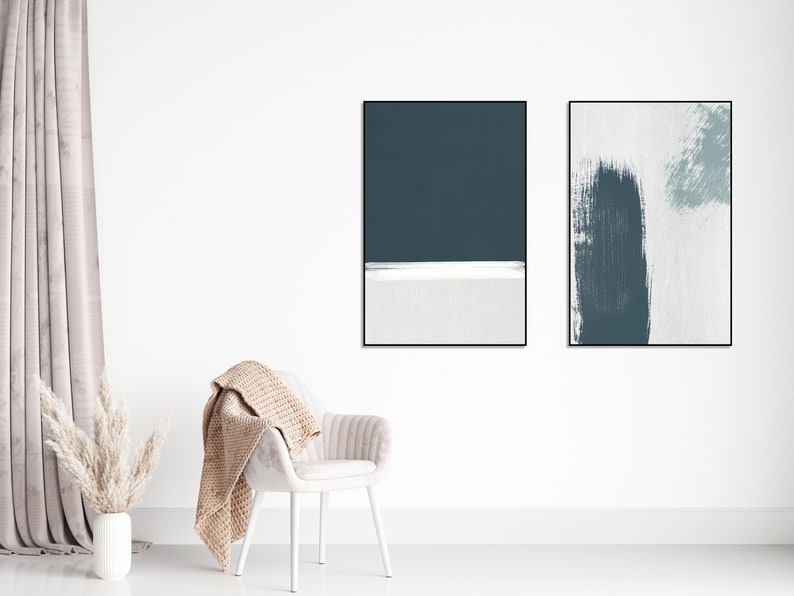 May include: Two framed abstract art prints with a white background and blue brushstrokes. The prints are hanging on a white wall above a white chair with a beige throw blanket.