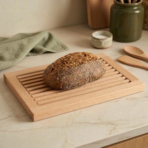 Puede incluir: Una tabla de cortar pan rectangular de madera clara con un diseño de superficie de listones. Una hogaza de pan artesanal de color marrón oscuro, con corteza crujiente y semillas y granos visibles, descansa sobre la tabla de madera.