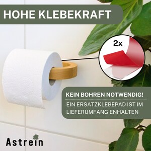 May include: A wooden toilet paper holder with a white roll of toilet paper. The holder is attached to a white tiled wall with a strong adhesive pad. The text "HOHE KLEBEKRAFT" is visible at the top of the image. The text "KEIN BOHREN NOTWENDIG! EIN ERSATZKLEBEPAD IST IM LIEFERUMFANG ENTHALTEN" is visible at the bottom of the image. The text "2x" is visible in a red circle.