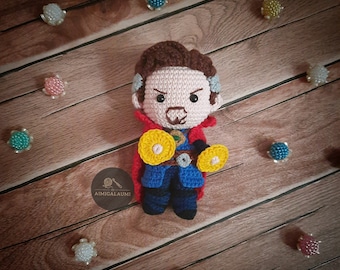 May include: A crocheted amigurumi doll of Doctor Strange from the Marvel Cinematic Universe. The doll is wearing a red cape, blue shirt, and holding two yellow amulets. The doll is sitting on a wooden surface.