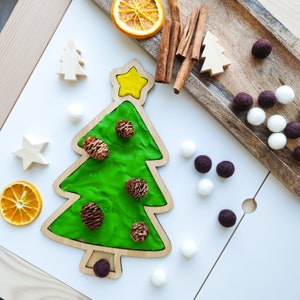 May include: A wooden Christmas tree cutout with green play dough inside. The tree is decorated with small brown pine cones and a yellow star on top. There are white and brown pom poms scattered around the tree.