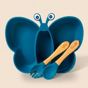 Peut inclure: Une assiette compartimentée en silicone bleu sarcelle en forme de papillon avec deux antennes et des yeux. Elle est accompagnée d'une fourchette et d'une cuillère assorties à manche en bois clair. La cuillère porte une étoile et le nom Camille.