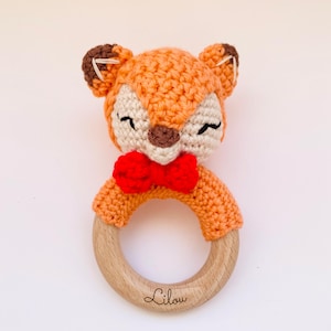 May include: A crocheted fox rattle with a red bow and a wooden ring. The ring is engraved with the name "Lilou".