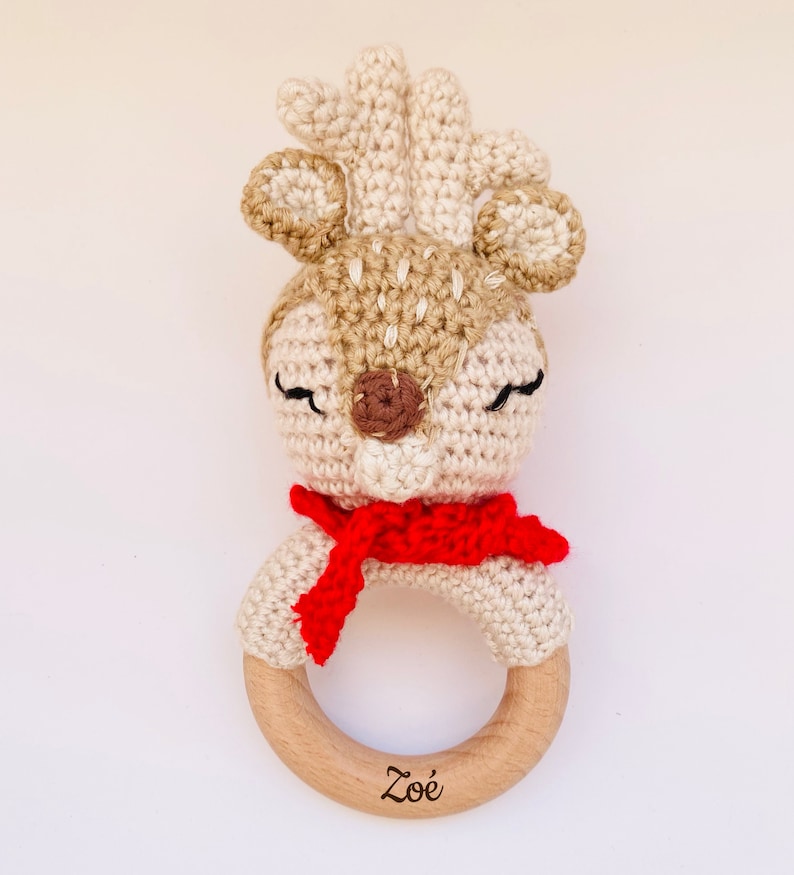 May include: A crocheted deer rattle with a red scarf and a wooden ring. The ring is engraved with the name "Zoe".