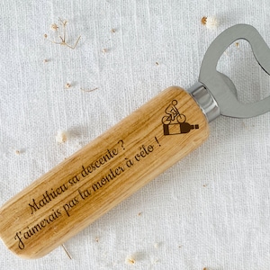 May include: Wooden bottle opener with a metal cap opener. The handle is engraved with the text "Mathieu sa descente? J'aimerais pas la monter &agrave; v&eacute;lo!" and a small image of a person riding a bicycle.