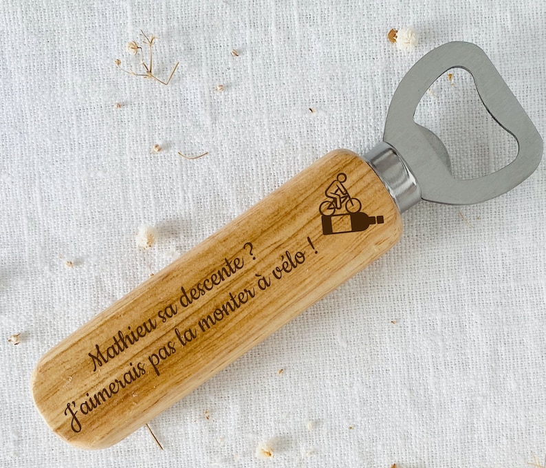 May include: Wooden bottle opener with a metal cap opener. The handle is engraved with the text "Mathieu sa descente? J'aimerais pas la monter &agrave; v&eacute;lo!" and a small image of a person riding a bicycle.