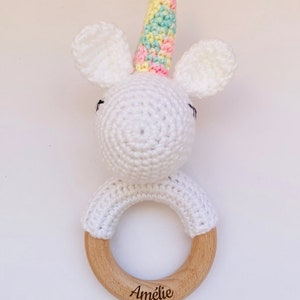May include: White crocheted unicorn rattle with a rainbow colored horn and wooden ring. The ring is engraved with the name "Amelie".