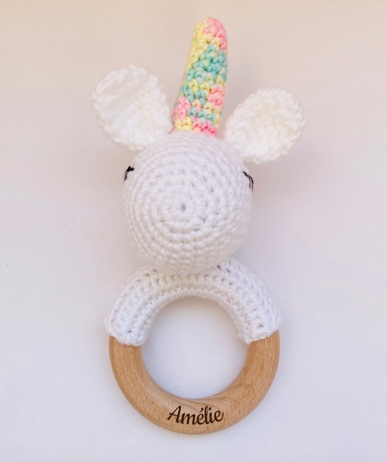 May include: White crocheted unicorn rattle with a rainbow colored horn and wooden ring. The ring is engraved with the name "Amelie".