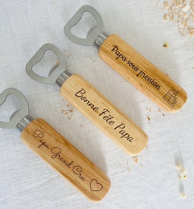 May include: Three wooden bottle openers with engraved messages. The first opener says "Papa sous pression" with a small coffee maker icon. The second opener says "Bonne F&ecirc;te Papa". The third opener says "Papa Grand Cru" with a heart icon.