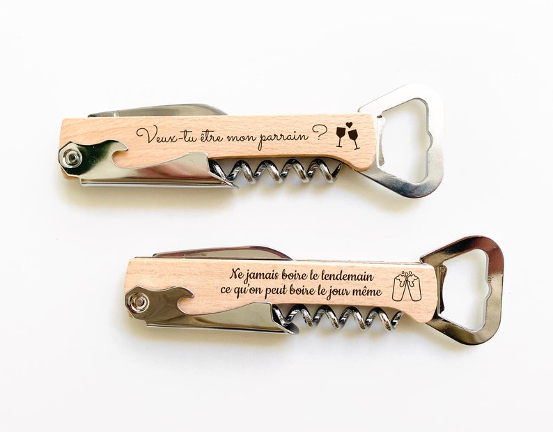 May include: Two wooden wine bottle openers with engraved sayings. The top opener says "Veux-tu &ecirc;tre mon parrain?" with a drawing of two wine glasses. The bottom opener says "Ne jamais boire le lendemain ce qu'on peut boire le jour m&ecirc;me" with a drawing of two wine glasses.
