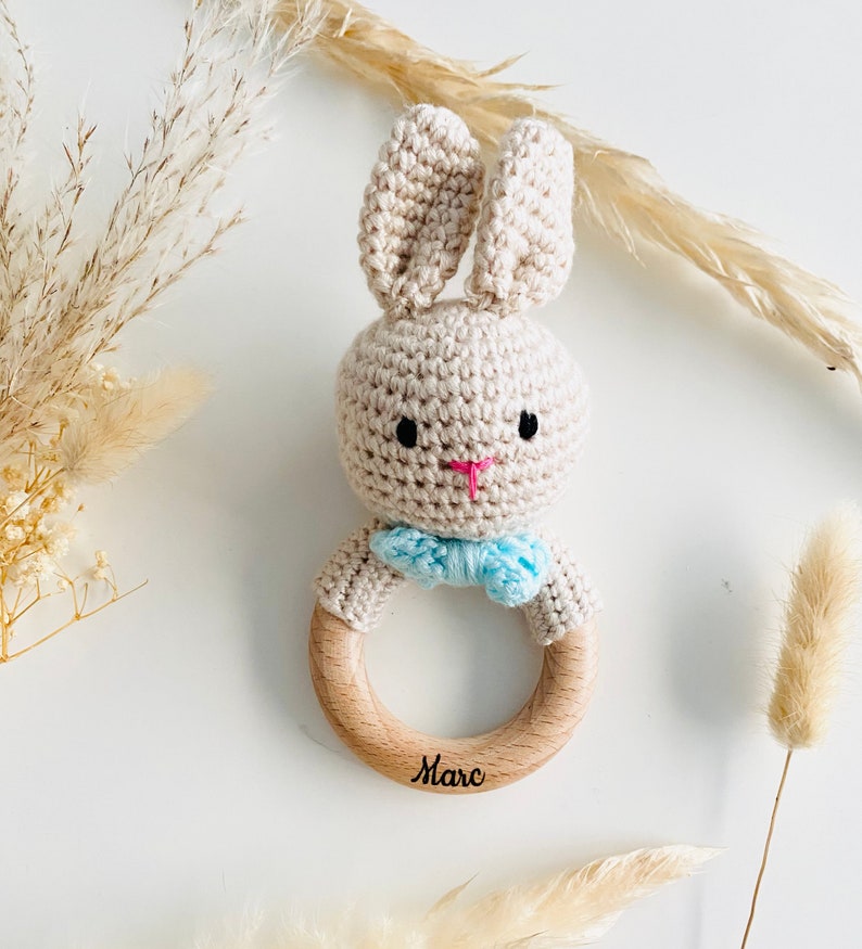 May include: A beige crocheted bunny rattle with blue bow tie attached to a wooden ring. The ring is engraved with the name "Marc".