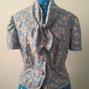 May include: A short-sleeved blouse featuring a blue, light blue, and beige botanical leaf and floral pattern. The garment has a high collar with a tied bow detail at the front, short puff sleeves, and a button-down front closure with matching fabric buttons.