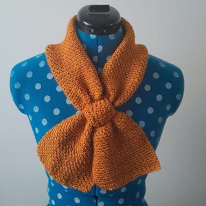 May include: An orange-colored, hand-knitted scarf is draped around a blue mannequin. The scarf is knotted at the front, showcasing its textured knit design. The scarf's warm hue contrasts with the mannequin's polka-dotted pattern, highlighting the handmade craftsmanship.