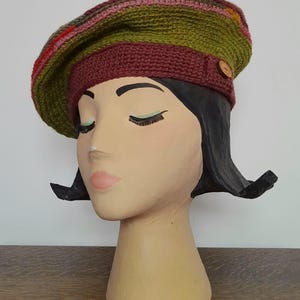 Puede incluir: Un gorro de punto de lana con un patrón de rayas verdes, marrones, rosas y rojas. El gorro tiene una banda marrón con un botón.